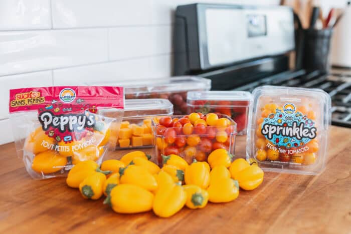 Containers of Sunset® Sprinkles tomatoes, Peppy's teeny tiny peppers, grapes, strawberries, and cheese cubes arranged on a kitchen counter next to a stove.