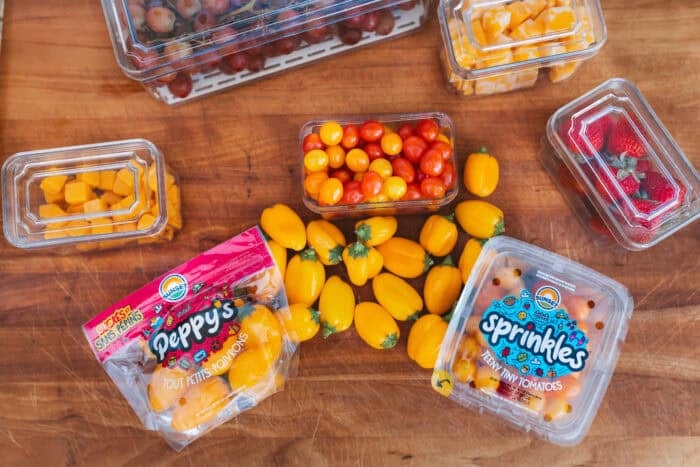 Top-down view of prepped produce and snacks including Sunset® Sprinkles tomatoes, Peppy's teeny tiny peppers, strawberries, grapes, cheese cubes, and tomatoes on a wooden surface in plastic containers.