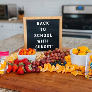 Back-to-school snack board on a kitchen island featuring Sunset® Sprinkles tomatoes, Peppy’s teeny tiny peppers, fruit, cheese cubes, and a letterboard sign reading “Back to School with SUNSET®.”
