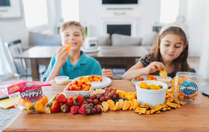 Two kids enjoy a colorful back-to-school snack board with Sunset® Sprinkles tomatoes, Peppy's teeny tiny peppers, strawberries, grapes, clementines, and crackers on a wooden board in a bright kitchen.