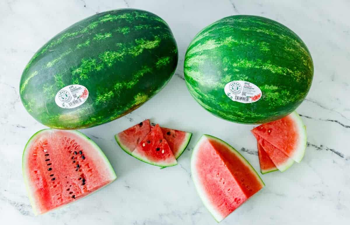 Two whole red watermelons - one seeded and one seedless -- next to sliced wedges on a marble surface, highlighting the visual difference between varieties.