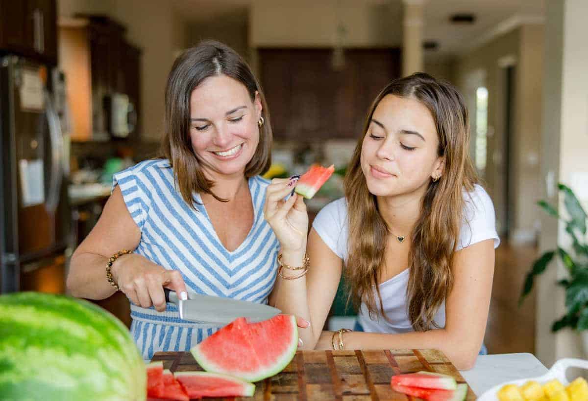 A mother slicing seedless watermelon while her daughter enjoys a slice, sharing a summer moment in the kitchen focused on healthy, seasonal snacking.
