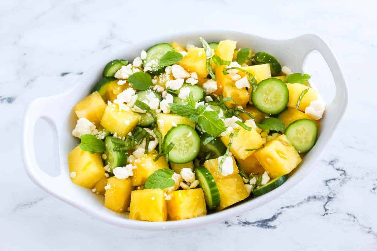 A vibrant summer salad made with cubed yellow watermelon, sliced cucumber, crumbled feta, and fresh herbs like mint and basil, served in a white dish for a refreshing side.