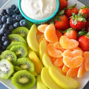 Cream cheese fruit dip in bowl plated with blueberries, kiwi, yellow apples, mandarins and strawberries.